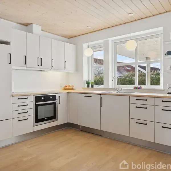 Køkken med hvide skabe og sort håndtag, ovn og vask. Rummeligt med træloft og trægulv. Store vinduer giver udsigt til have. Hylder med kopper og en kaffemaskine.