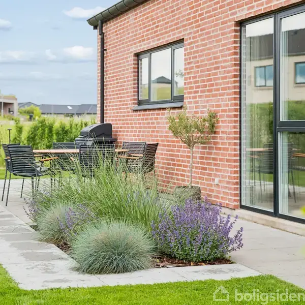En terrasse foran et rødt murstenshus med glasdør og vindue. Bord og stole står ved siden af planter og lavendel langs en flisebelagt sti, omgivet af en grøn have.