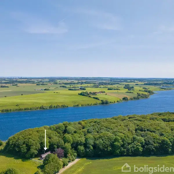 Hus omgivet af grønne træer ved skovkant, pil peger mod det. En bred flod løber forbi, med vidtstrakte marker på begge sider under en klar himmel.