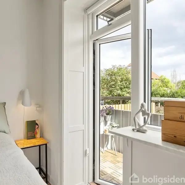 Soveværelse viser et åbent vindue med udsigt til en balkon. Bøger og en skulptur pryder vindueskarmen, mens en lampe og bog er på bordet ved siden af sengen.