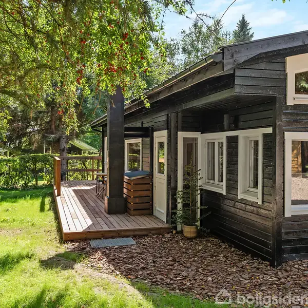 Sortmalet træhus med store vinduer og en træterrasse. Omgivet af grønne træer og græsdækket have, skaber en fredfyldt atmosfære. Søjler understøtter tagudhæng over terrassen.