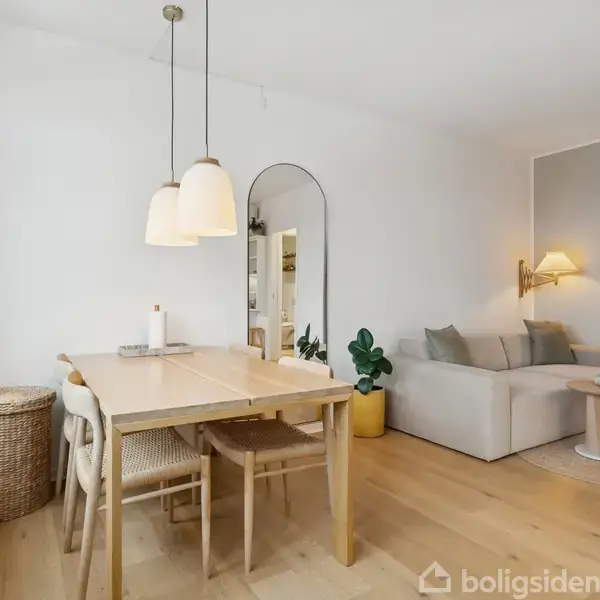 Et spisebord med to stole står i hjørnet af et lyst rum med parketgulv. Over bordet hænger to lamper. En sofa er placeret ved siden af en væg med en lampe.