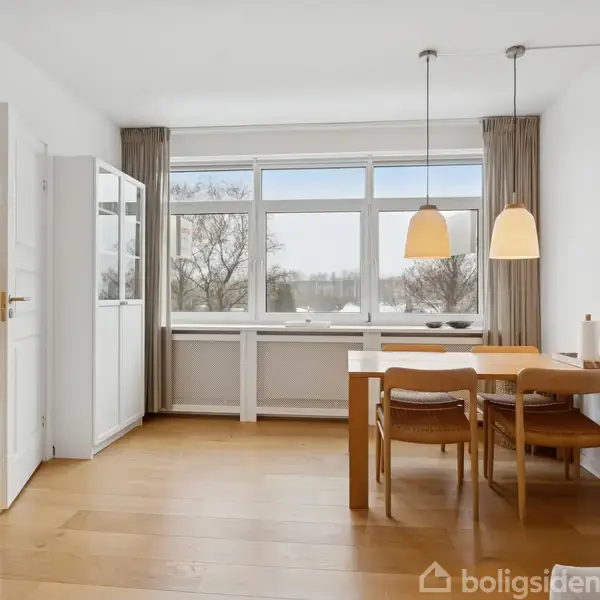 En spisestue med et træbord og fire stole er centralt placeret under to hængende lamper. Rummet har trægulv, store vinduer med naturudsigt, og hvide vægge.