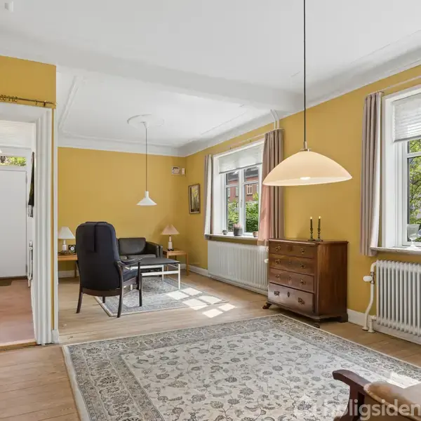 Et hyggeligt stueområde med gule vægge og store vinduer. En sort stol og bord står ved siden af en kommode. Et tæppe pryder gulvet, og lamper hænger fra loftet.