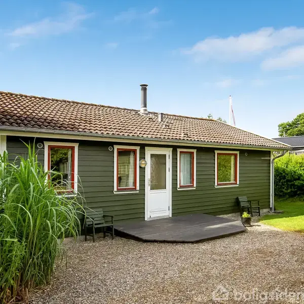 Grønt træhus med terracotta-tag står stille på en grusbelagt gårdsplads omkranset af frodig have. Skilt viser ingen tekst.