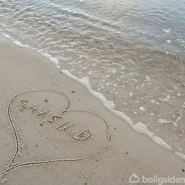 Et hjerte er tegnet i sandet, indeholdende teksten "SALGSILD", nær vandkanten på en strand, hvor små bølger ruller ind mod kysten.
