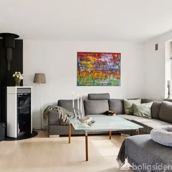 Gråt hjørnesofa-arrangement med glasbord står i en lys stue. Farverig kunst pryder væggen. Brændeovn, stående lampe og vindue med udsigt til grøn have skaber hyggelig atmosfære.