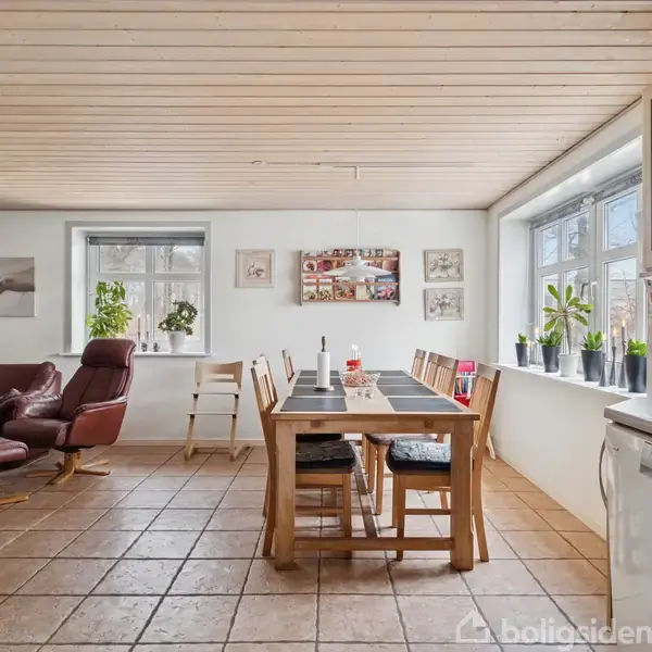 Et spisestuebord står centralt i et lyst rum, omgivet af træstole. Foran vinduerne til højre står planter. En sofa- og stolgruppe er til venstre med billeder på væggene.