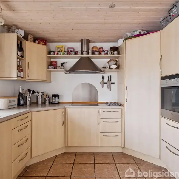 Køkken med lyse træskabe. Hætte over komfur, omgivet af køkkenredskaber og krydderier. Indbygget ovn og køleskab til højre. Keramisk flisegulv, træloft, hvid væg.