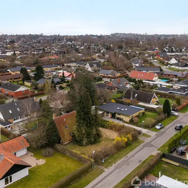 Parcelhuse omgivet af grønne haver set fra oven, beliggende i en forstadskvarter med snoede veje og tæt beplantning.