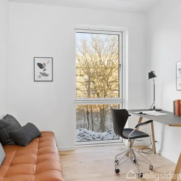 En minimalistisk arbejdsplads med et skrivebord og stol står ved et vindue med udsigt til træer. Rummet har hvidmalede vægge, en brun sofa og to billeder på væggen.