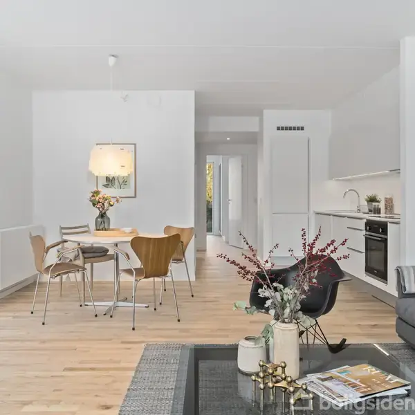 Moderne stue med gråt sofabord og sofa, dekoreret med plante. Rundt spisebord med stole står nær åbent køkken. Store vinduer giver lys, hvilket skaber en lys og indbydende atmosfære.