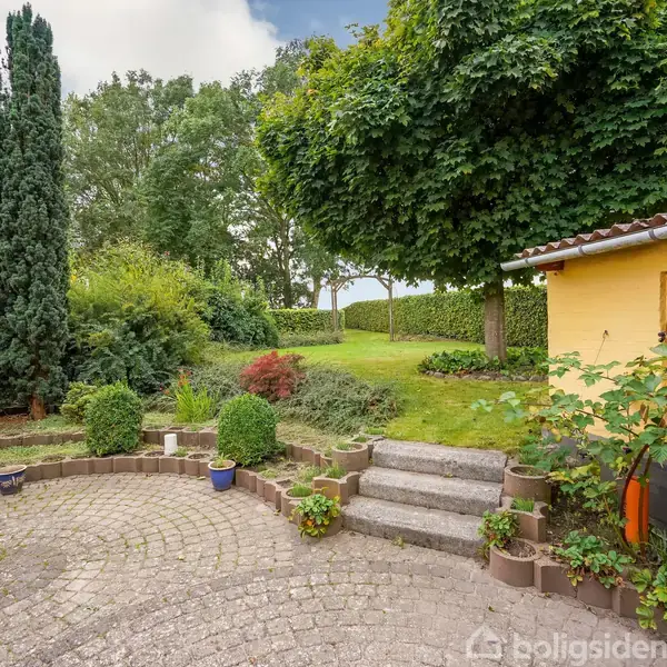 Terrasse med gul bygning omgivet af grønne haver. Stentrin fører op til en græsplæne med store buske og høje træer i baggrunden, der skaber en frodig, privat atmosfære.