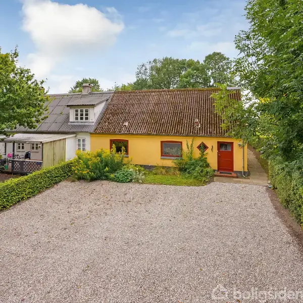 Et gult hus med en brun tag og en rød dør står ved en grusbelagt indkørsel, omkranset af grønne buske og træer, med en blå himmel i baggrunden.