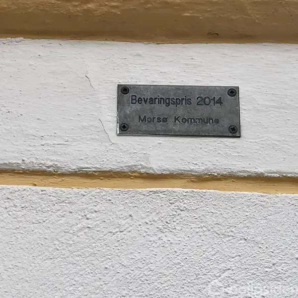 Metalplade er fastgjort til væg med teksten: "Bevaringspris 2014 Morsø Kommune" i en murstensstruktur. Pladen angiver bevaringsprisanerkendelse. Omgivelserne viser en hvid og gul facade.