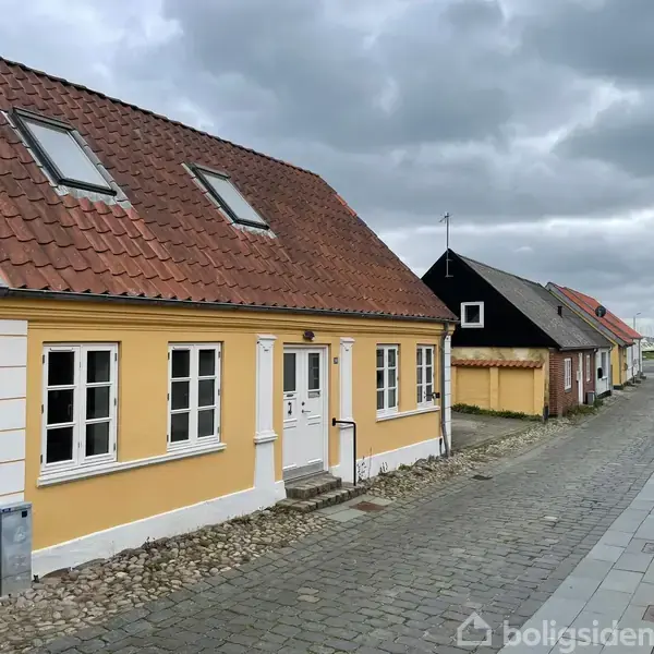 Et gult hus med hvide vinduer og rødt tag står langs en brostensbelagt gade i et lille nabolag. Skydække dækker himlen og flere huse ses langs gaden.