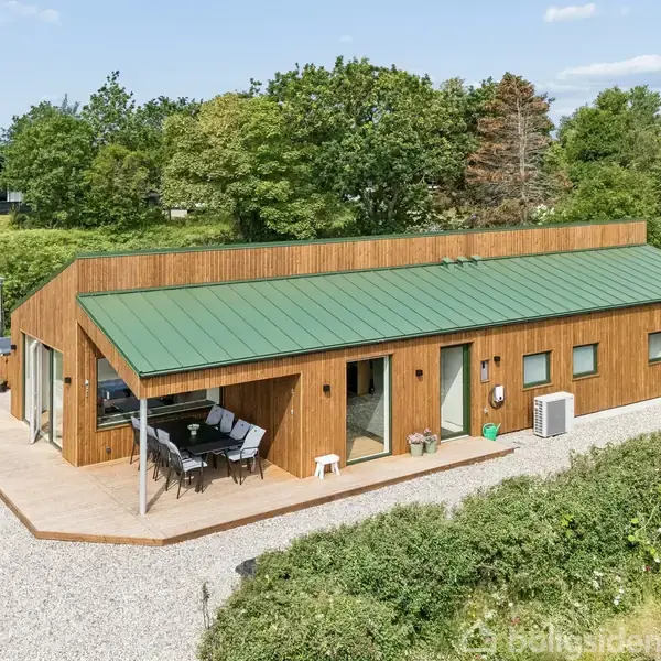 Et moderne træhus med grønt tag står omgivet af en frodig have. En terrasse med havemøbler ses foran huset. Omkringliggende træer og buske indrammer ejendommen.