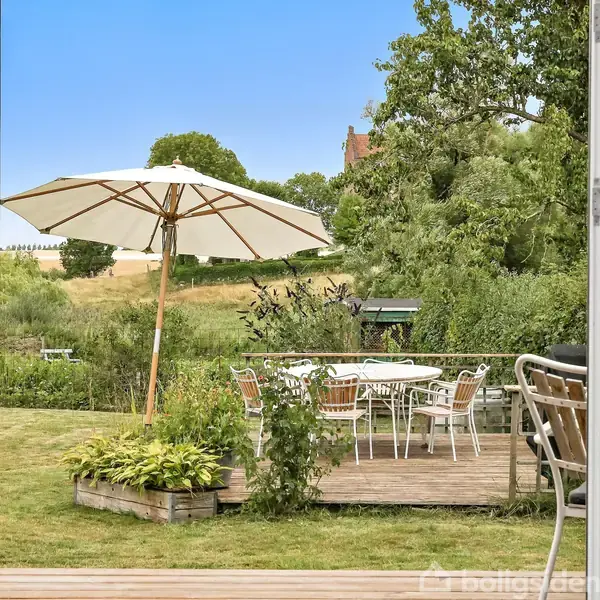 Udendørs terrasse med hvidt parasol over et bord og stole, omgivet af grønne planter. I baggrunden ses en stor have med træer og en bygning i det fjerne.