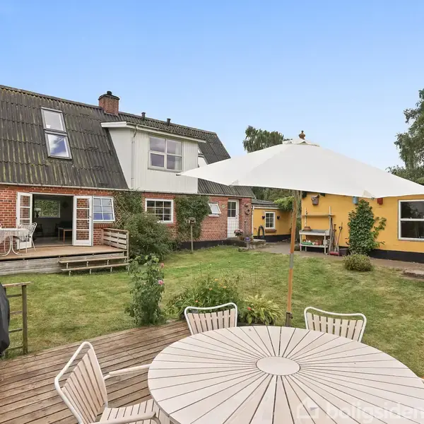 Rundt havebord med parasol står på en træterrasse. I baggrunden ses et hus med røde teglsten og sort tag, samt en gul tilbygning med store vinduer i grøn have.