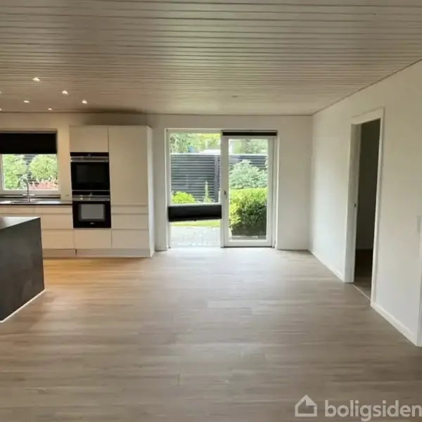 Køkken-alrum med lyst trægulv og hvidt loft. Køkkenet har hvide skabe, indbygget ovn, og stor vindue/dør mod have, der lukker naturligt lys ind.