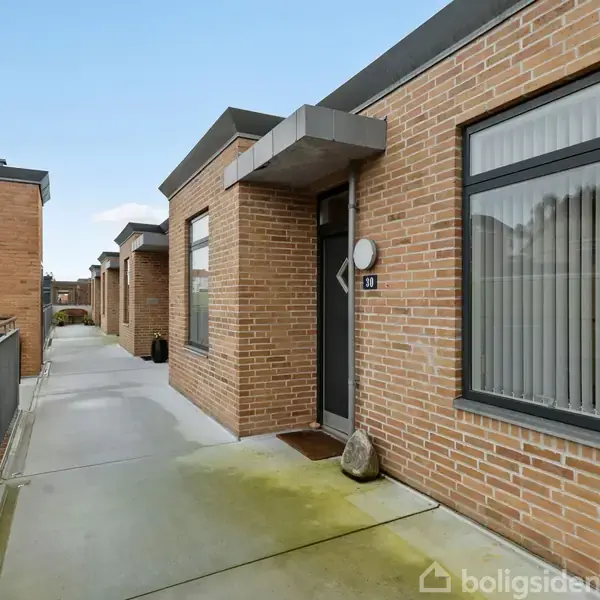 En murstensbygning med store vinduer står langs en gangsti. Nummeret "21" ses ved indgangen, og gangstien har rækværk langs siden. Himlen er let skyet.