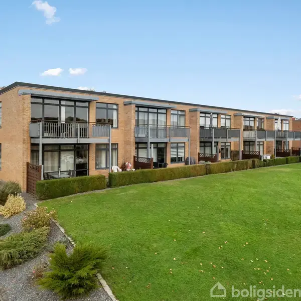 En række moderne, to-etagers murstenslejligheder med balkoner står i en grøn plæne omgivet af dekorative buske og en grussti, under en klar blå himmel med få skyer.