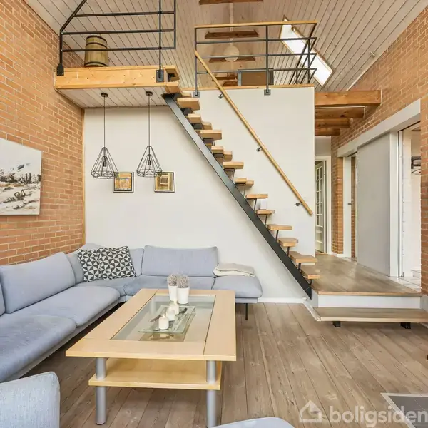 Stue med lys blå hjørnesofa og sofabord, placeret under en moderne trappe. Væggene er af røde mursten, og der hænger kunstværker. Ovenpå er der en hems med trægulv.