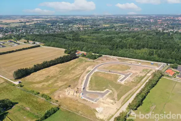 Stykke land under udvikling med anlagte veje, omkranset af skov og marker, set fra luften. I baggrunden ses en by i horisonten.
