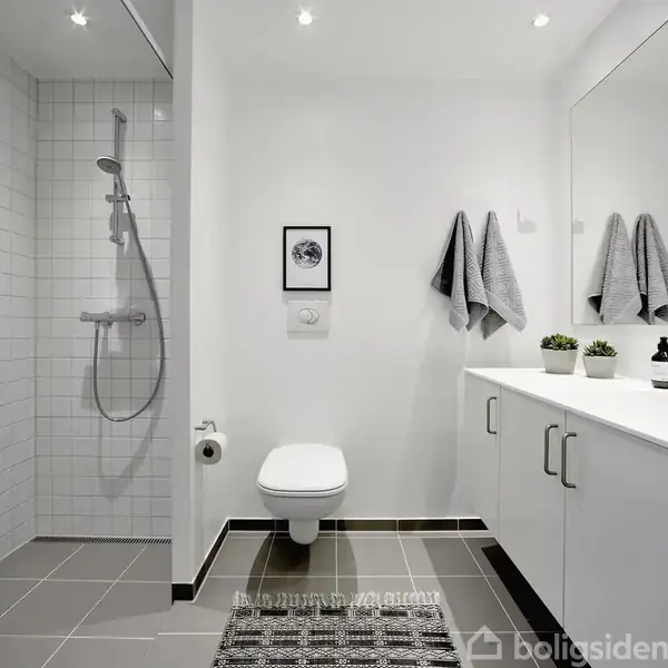 Toilet hænger på væggen i et moderne badeværelse. Ved siden af er der en bruseniche med en bruser, og til højre er der et langt hvidt skab og spejl over vasken.
