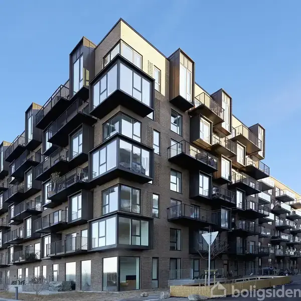 Moderne lejlighedsbygning med mange altaner og store vinduespartier beliggende i et byområde om dagen, omgivet af en blå himmel.