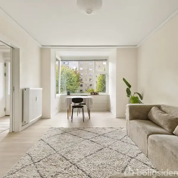 Et lyst værelse med en beige sofa står stille, mens et bord og en stol er placeret foran et stort vindue med udsigt til træer og bygninger. En grøn plante står ved siden af sofaen.