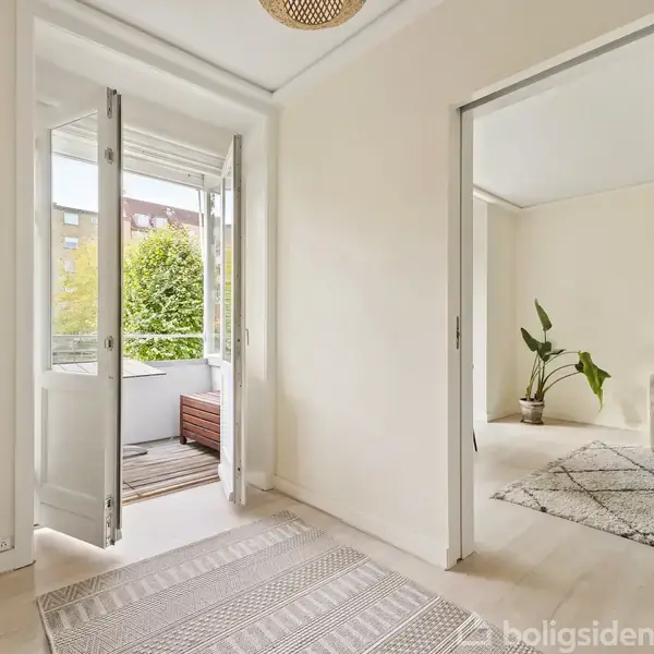 Døråbning til altan med hvide døre; inde i lejligheden ses en stue med beige sofa og tæppe. Altanen har trædæk og udsigt til grønne træer og bygninger.