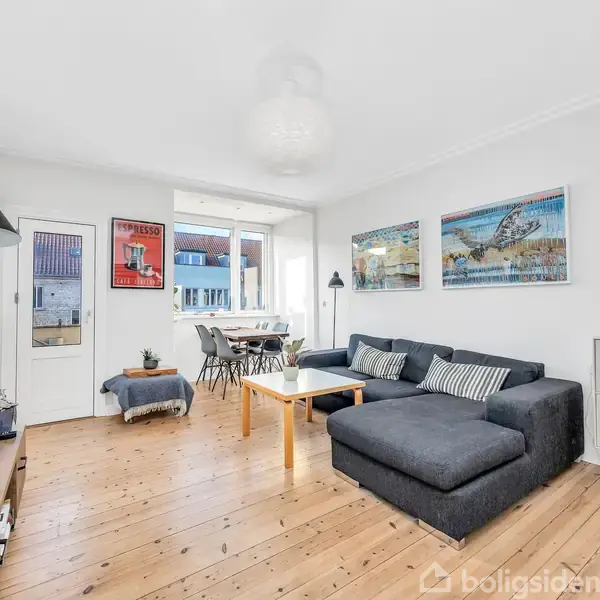 Stue med en stor grå hjørnesofa foran et TV, placeret på et trægulv. Vindue i baggrunden med spisebord og stole. Vægkunst og lamper som dekorationer.