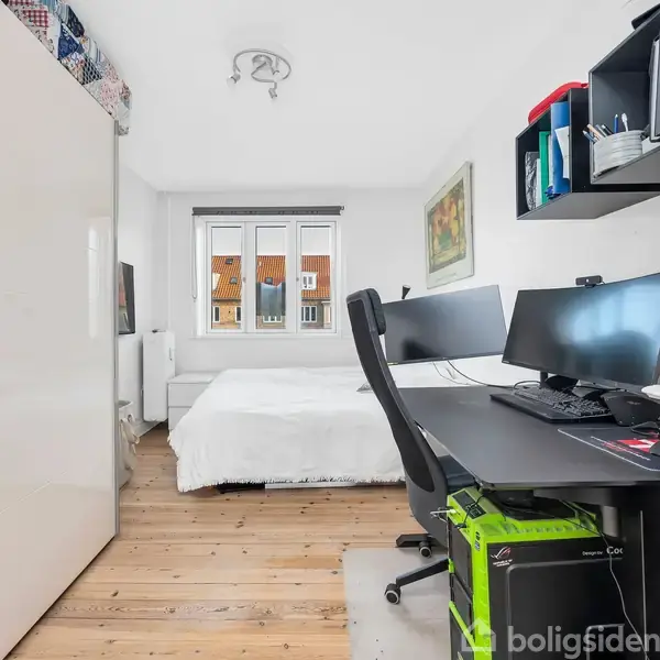 Et moderne kontor kombineret med soveværelse. Computeren står på et sort skrivebord med to skærme. Ved siden af er en seng op ad væggen. Reol over skrivebordet.