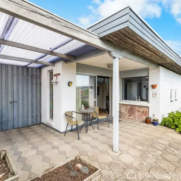 Overdækket terrasse med bord og stole; tilstødende hvid bygning med store vinduer og trægavl; flisebelagt gulv omgivet af haver i et solrigt udemiljø.