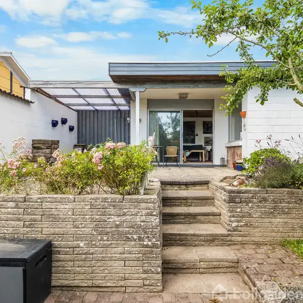 En lille terrasse er forbundet til et hvidt hus. Trin og blomsterbede fører op til terrassen. Dækket sideområde til venstre, græsplæne til højre.