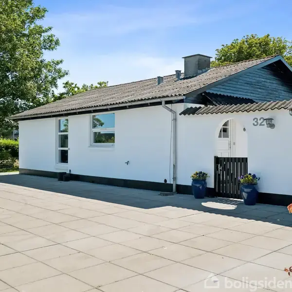 Et hvidt hus med sorte detaljer står stille på en flisebelagt grund. Der er to blå krukker ved indgangen flankeret af en grøn have. Nummer 32 ses ved døren.