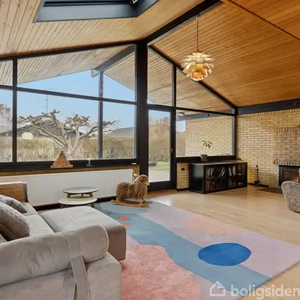 Stue med stor hjørnesofa og tæppe i farverigt mønster. Store vinduer giver udsigt til have og lysindfald. Loftet er træbeklædt, og der hænger en unik loftslampe. Pejs i hjørnet.