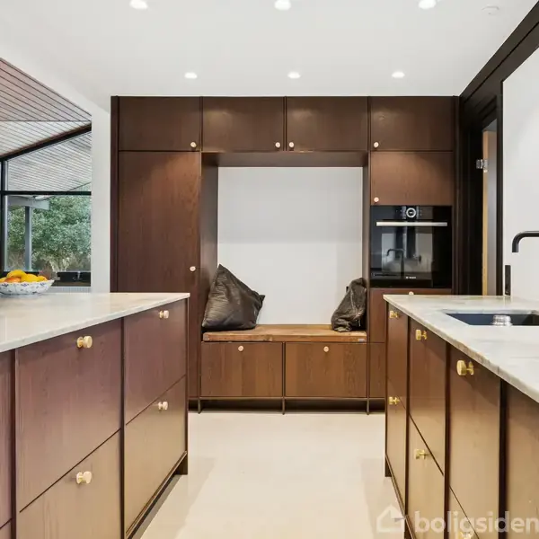 Køkken med mørke træskabe dominerer, mens det centrale øområde har en skål med frugt. Baggrunden viser store vinduer og bestemmelsesplads med puder i et moderne, lyst rum.