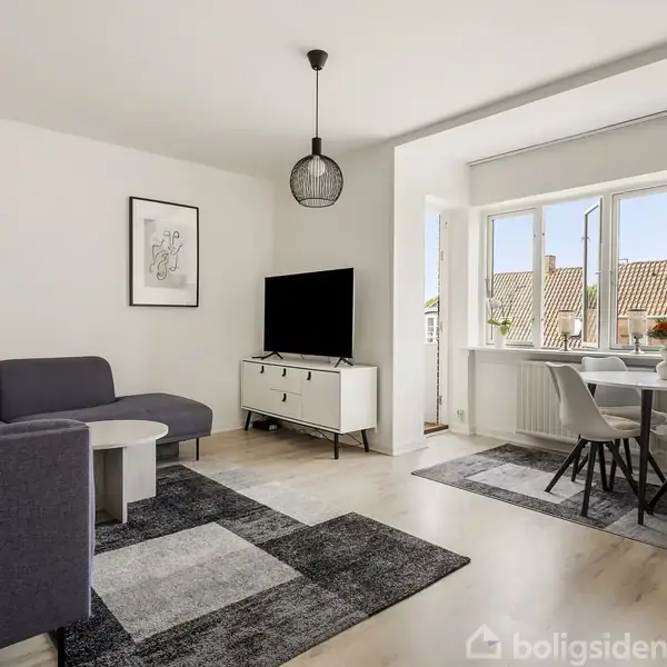 Stue med mørkegrå sofa og sofabord, hvor et TV står på et hvidt skab. Ved vinduet står et spisebord med fire stole. Rummelig, lyst interiør med tæpper og lampe.