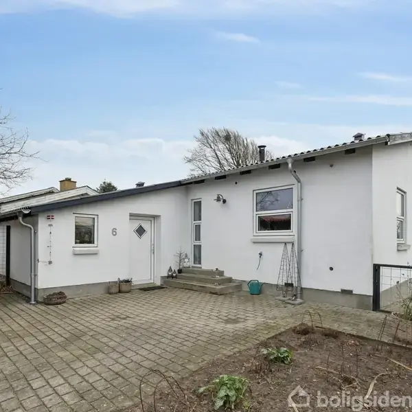 Hvidt hus med nummer 6, fliseterrasse og lille trappe, omgivet af buske og træer i en have. Himmel er blå med få skyer.