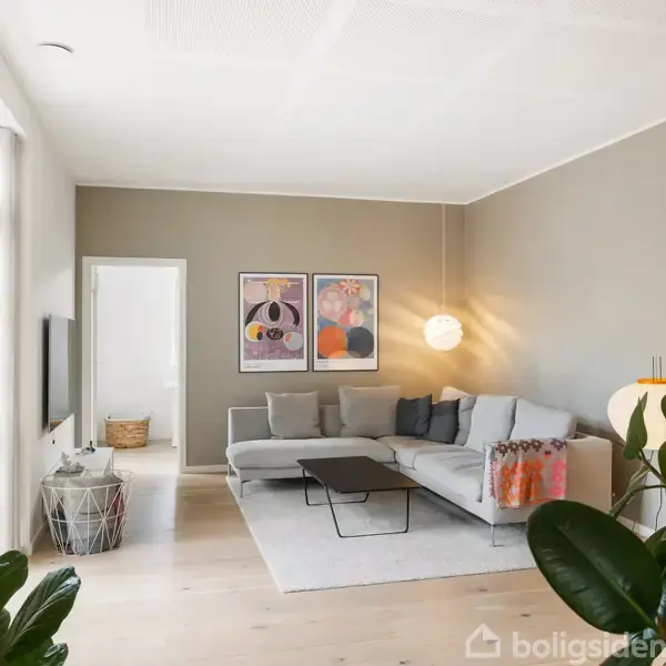 En lys stue med en stor grå sofa står langs væggen. To farverige plakater hænger over sofaen, og en rund lampe lyser rummet. Planter og kunst dekorerer hjørnerne.