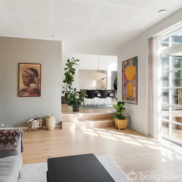 Sofa i en moderne stue, belyst af naturligt lys gennem store vinduer. Stuen fører til en spisestue med et stort bord, planter og kunst på væggene. En plakat viser en kvinde.