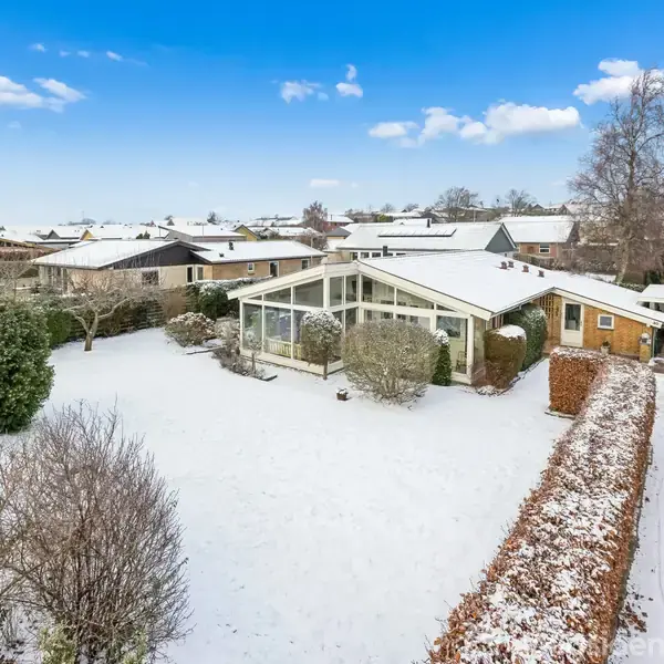 Hus beliggende i et snedækket nabolag med en stor have omgivet af træer og buske. En indkørsel fører til en carport på højre side. Solrig himmel med spredte skyer.