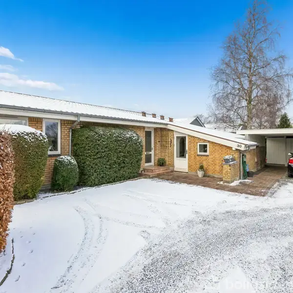 Et gult murstenshus med sne på taget står i en indkørsel. En bil er parkeret i en carport til højre, omgivet af snedækkede buske og træer.