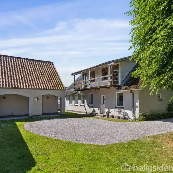 En hvid bygning med rødt tegltag står stille i en gård med grusbelægning, omgivet af grøn bevoksning og en træ i højre side under en blå himmel.