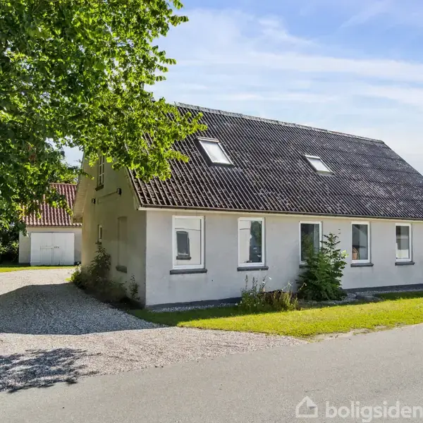 Et hvidt hus med sorte tagsten står ved en vej. Huset har flere vinduer og en grusvej ved siden af fører til en garage. Grønt græs omgiver stedet.