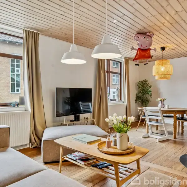 Sofa står i en stue med grå polstring. Over bordet hænger loftslamper, og en Peppa Gris ballon svæver. I baggrunden er spisebord og vinduer med lyse gardiner.