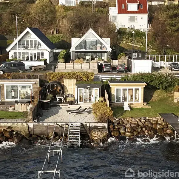 To huse med store vinduespartier står ved en skråning ved vandet. Foran dem er en terrasse med havemøbler på en grusflade omgivet af grønne buske og store sten.
