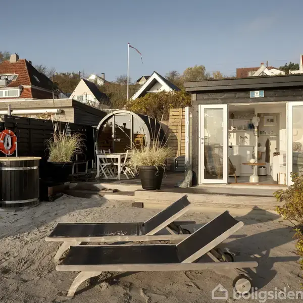 En lille, moderne strandhytte står med åbne døre, omkranset af sand, terrassemøbler og en spabad. I baggrunden er der flere hvide huse på en skråning.